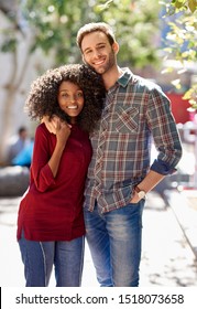 Portrait Of A Diverse Young Couple Affectionately Standing Arm In Arm Together On A City Sidewalk On A Sunny Day