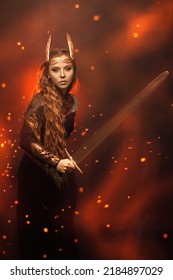 Portrait Of Divine Female Warrior Valkyrie Standing With A Sword In Her Hand. Epic Fantasy. Scandinavian Mythology. Dark Background With Smoke And Red Light.