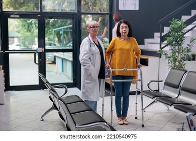 Portrait Of Doctor Doing Physical Therapy Exam With Injured Patient, Supporting With Walking Frame To Recover After Leg Fracture Injury. Asian Woman Attending Medical Physiotherapy Consultation.