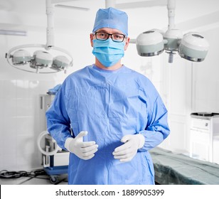 Portrait of doctor in glasses looking at camera while standing in operating room. Man surgeon in sterile gloves, blue surgical uniform and protective face mask, ready for plastic surgery in clinic - Shutterstock ID 1889905399