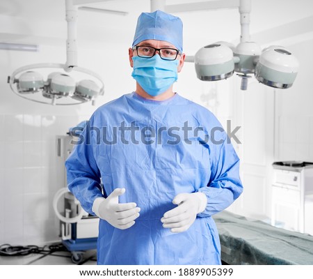 Portrait of doctor in glasses looking at camera while standing in operating room. Man surgeon in sterile gloves, blue surgical uniform and protective face mask, ready for plastic surgery in clinic