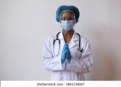 Portrait Of A Doctor In Lab Coat With Face Mask, Face Shield,  And Hair Ppe With Gloved Hands Together As If Saying Please Or Praying