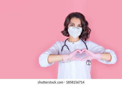 Portrait of a doctor in a medical protective mask with a stethoscope and pink nitrile gloves. Hands showing a healthy heart symbol of love, cardiology. Isolate on a pink background. Copy space