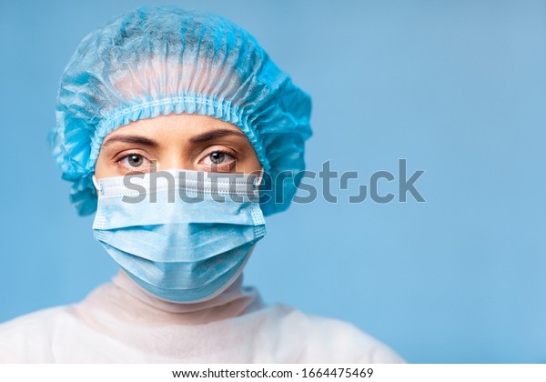 Portrait of a doctor, a young woman in a protective\
medical mask on her face and a cap on her head. looking seriously\
into the frame. on a blue background. surgeon. ambulance paramedic.\
copy space