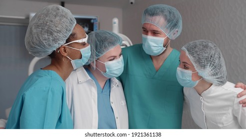 Portrait Of Doctors In Uniform And Safety Mask Hugging Posing At Camera In Dental Office. Diverse Team Of Dentists Embracing And Smiling Working In Modern Clinic