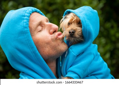 Portrait Of Dog Owner Kissing A Furry Friend In Matching Blue Hoodies Outdoors In Bright Green Park Background