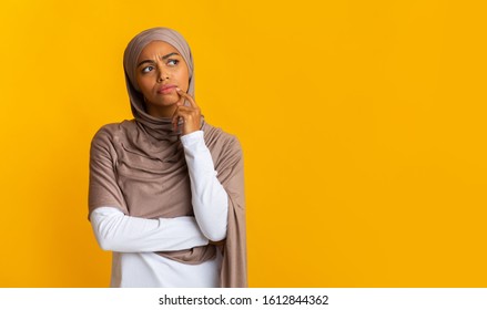 Portrait of doubtful afro muslim woman in headscarf touching chin, overthinking options and looking away over yellow background