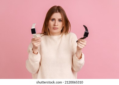 Portrait of doubtful woman holding two smart watches in hands, having difficulty what wristwatch to choose, looking at camera, wearing white sweater. Indoor studio shot isolated on pink background. - Powered by Shutterstock