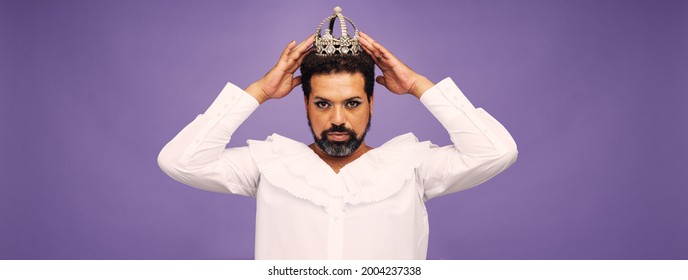 Portrait Of A Drag Queen Posing On Purple Background. Bearded Man Wearing Makeup And Crown Looking At Camera.