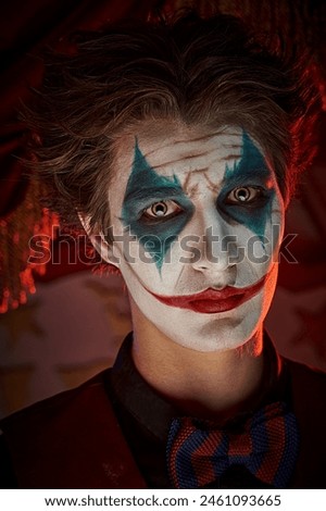 Portrait of a dramatic young man with clown, joker makeup looking sad on a dark circus background. Halloween. Drama, horror.
