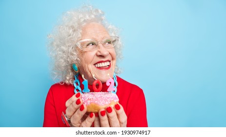 Portrait of dreamy cheerful curly elderly woman smiles broadly concentrated aside thinks about wish before blowing candles wears red jumper jewelery bright makeup holds tasty glazed doughnut - Powered by Shutterstock