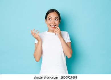 Portrait Of Dreamy Clueless Cute Asian Girl In White T-shirt Biting Fingernail And Looking Thoughtful Up, Thinking What To Do, Making Decision, Standing Blue Background And Pondering