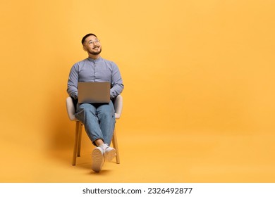 Portrait Of Dreamy Smiling Asian Man With Laptop Sitting In Chair And Looking Up At Copy Space, Happy Millennial Male Using PC Computer, Working Or Study Online Over Yellow Studio Background