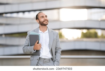 Portrait Of Dreamy Young Businessman With Laptop In Hand Standing Outdoors And Looking Aside At Copy Space, Smiling Handsome Male Entrepreneur In Suit Thinking About Business Plan, Copy Space - Powered by Shutterstock