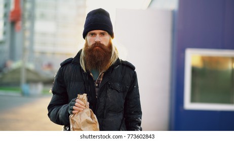 Portrait of Drunk homeless man with drink alcohol from paper bag while standing on the city street and looking at camera