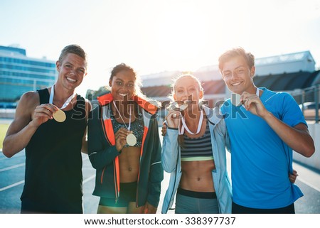 Portrait of ecstatic young runners showing medals. Young men and women looking excited after winner a running race. Team of multiracial athletes in stadium.