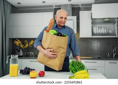 Portrait of elder happy man holding paper bag with provision. Bread and vegetables in paper pack in grandfather hands standing at table of home kitchen. - Shutterstock ID 2255692861