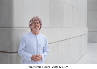 Portrait of elderly Arab man standing confidently wearing traditional kandora attire and headscarf, posing against a modern, minimalist building with white, clean lines neutral tones and copy space. - Powered by Shutterstock