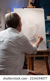 Portrait Of Elderly Artist Sketching Still Life Using Sharp Pencil On Paper Sketching Vase Model In Home Art Studio. Over Shoulder View Of Older Woman Drawing Orginal Masterpiece In Workshop.