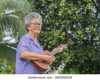 Portrait of an elderly Asian woman with short gray hair playing the ukulele while standing in a garden. Enjoy life after retiring. Concept of aged people and relaxation.