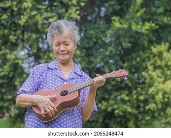 Portrait of an elderly Asian woman with short gray hair playing the ukulele with a smile while standing in a garden. Enjoy life after retiring. Concept of aged people and relaxation.