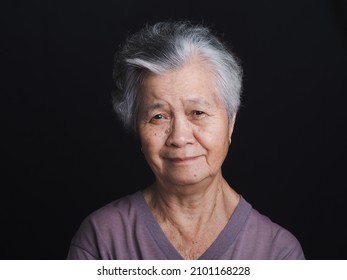 Portrait Of An Elderly Asian Woman With Short Gray Hair Looking At The Camera With A Smile While Standing On Black Background In The Studio. Space For Text. Aged People And Health Care Concept