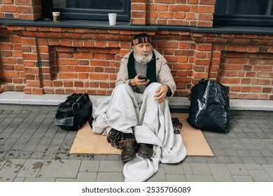 Portrait of elderly beggar man using smartphone sitting on cold winter street, wrapped in blanket for warmth, surrounded by meager belongings. Older tramp seek work and government assistance.