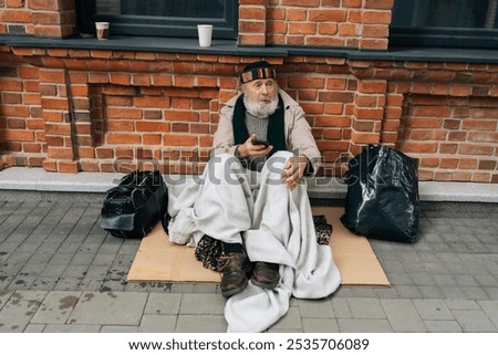 Portrait of elderly beggar man using smartphone sitting on cold winter street, wrapped in blanket for warmth, surrounded by meager belongings. Older tramp seek work and government assistance.