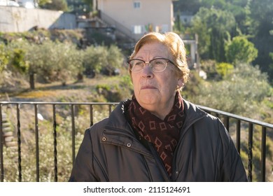 Portrait of an elderly Caucasian woman with glasses, with a serious and worried expression, wrapped in a coat, with an unfocused natural background.