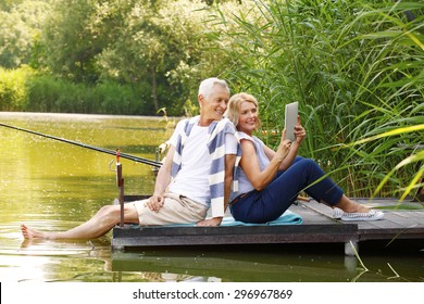 Portrait of elderly couple relaxing at nature. Senior woman and old man sitting back to back on the pier at lakeside while making self portrait with digital tablet.  - Powered by Shutterstock