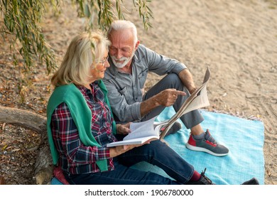 Portrait Of An Elderly Couple Sitting And Reading A Newspaper At The Riverside Beach.