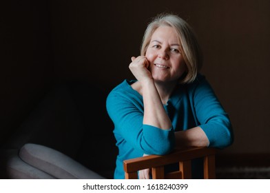 Portrait Of An Elderly Happy Well-groomed Beautiful Woman Blonde Pensioner 50-60 Years Old With Light Hair On A Dark Brown Background Of The House. Close-up, Space For Text