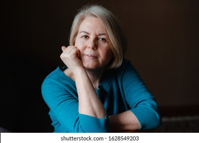 Portrait Of An Elderly Happy Well-groomed Beautiful Woman Blonde Pensioner 50-60 Years Old With Light Hair On A Dark Brown Background Of The House. Close-up, Space For Text