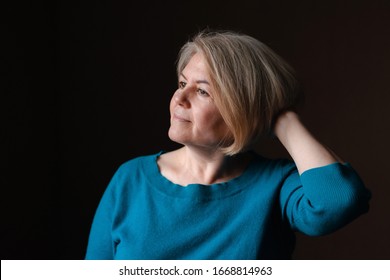 Portrait Of An Elderly Happy Well-groomed Beautiful Woman Blonde Pensioner 50-60 Years Old With Light Hair On A Dark Brown Background Of The House. Close-up, Space For Text