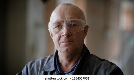 Portrait of elderly industrial worker on plant with blured background. Old man wearing transparent protective glasses and uniform inside of industrial plant. - Powered by Shutterstock