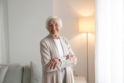 Portrait Of Elderly Lady In Beige Cardigan Alone At Home. Senior Woman 86 Years Of Age Standing By The Window In Her Apartment. Background, Copy Space, Close Up.