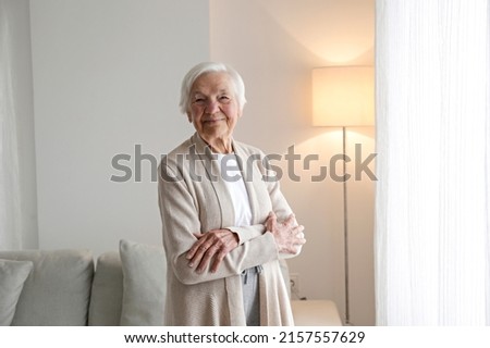 Portrait of elderly lady in beige cardigan alone at home. Senior woman 86 years of age standing by the window in her apartment. Background, copy space, close up.