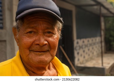 Portrait Of An Elderly Latin Man With A Cap Smiling In Front Of His House In A Poor And Rural Community In Latin America.