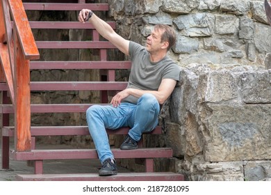 Portrait Of An Elderly Man 45-50 Years Old, Taking A Selfie On His Phone, Sitting On The Steps Against The Background Of An Old Stone Wall.