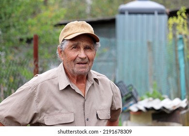 Portrait Of Elderly Man In Baseball Cap And Short Sleeve Shirt Standing In Rural Yard. Life In Village, Old Age