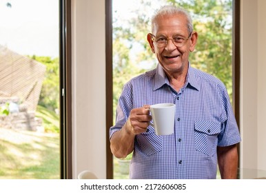 Portrait of an elderly man drinking coffee in front of a window where you can see the backyard of her house.