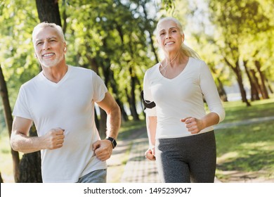 Portrait Of Elderly Man And Old Woman In Headphones Jogging Together Outside In Park