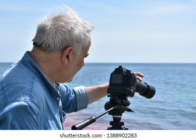 Portrait Of An Elderly Man Photographer By The Sea. Senior Adult Shooting Video With DSLR Camera With Telephoto Lens On Tripod.