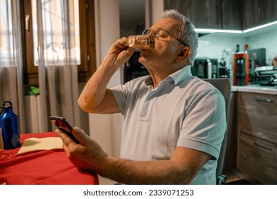 Portrait of elderly man wearing glasses and casual clothes holding mobile mobile phone, sitting in kitchen drinking coffee, breakfast, morning routine. Senior man taking break. Online technology - Powered by Shutterstock