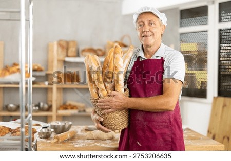 Portrait of elderly positive baker with bread and baguettes in hands in kitchen