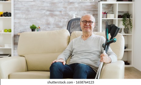 Portrait of elderly retired man smiling to the camera in cozy nursing home, he has crutches next to the sofa