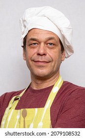 Portrait Of An Elderly Self-confident Male Chef 45-50 Years Old In A White Cap And Apron On A Light Background With Crossed Arms And Looking Into The Camera.