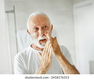 Portrait of an elderly senior man applaying a cream on his face in front of a mirror in bathroom, skincare, beauty and vitality concepts - Powered by Shutterstock