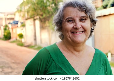 Portrait Of Elderly, Senior, Mature Woman Full Of Energy, Fun In Front Of Home Smiling Happy And Confident. Natural Light, Sunlight. Looking At A Camera Seen From The Front. Real Person