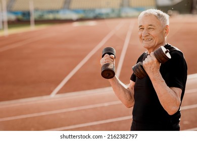 Portrait Of An Elderly Smiling Man In Black Sportswear. Senior Man Exercising With Dumbbells At The Stadium. Sport And Lifestyle Content. High Quality Photo